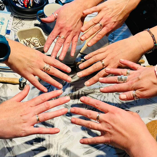 Multiple hands with rings reaching towards each other on a table with jewellery tools. 